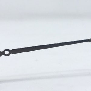 May include: A black metal clock hand with a slender, rectangular body and a circular opening near the base. The hand tapers to a point at the end, suggesting a time-telling function. The object is set against a plain white background.