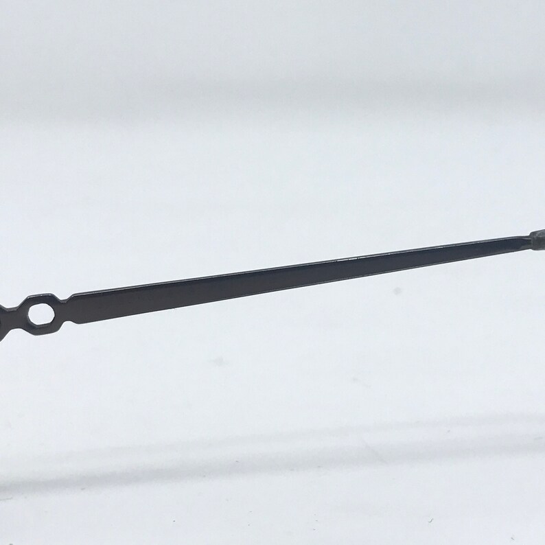 May include: A black metal clock hand with a slender, rectangular body and a circular opening near the base. The hand tapers to a point at the end, suggesting a time-telling function. The object is set against a plain white background.