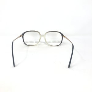 May include: Clear eyeglasses with a gradient gray and clear frame. The glasses have gold-colored arms and dark blue earpieces. The lenses are clear, and the overall style is vintage.