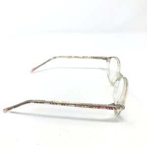 May include: Clear eyeglasses with round lenses and patterned temples. The temples feature a repeating geometric design in shades of red, pink, and gold. The frames are a transparent, light color.