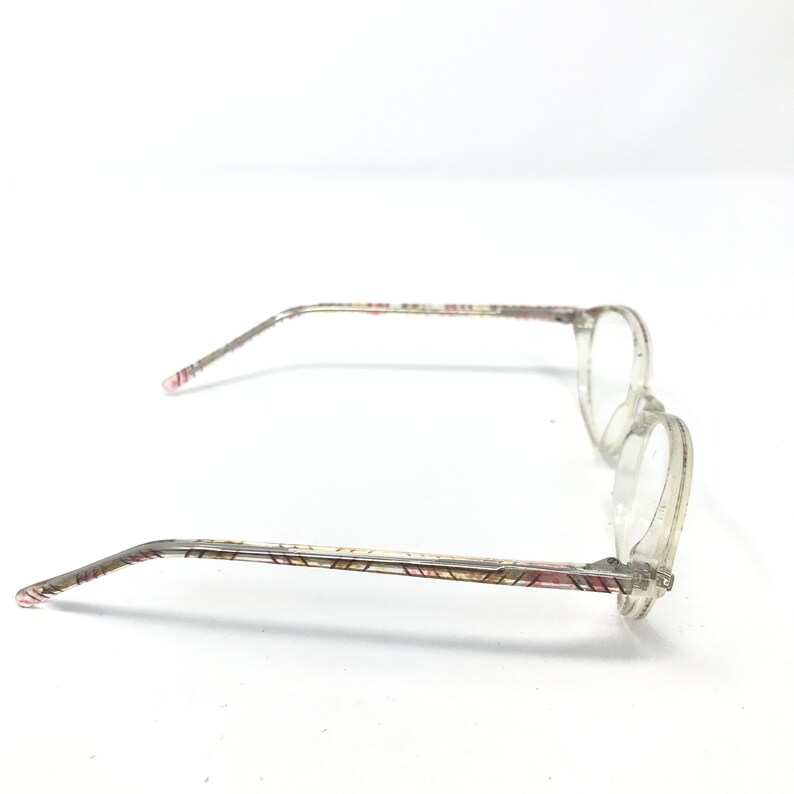 May include: Clear eyeglasses with round lenses and patterned temples. The temples feature a repeating geometric design in shades of red, pink, and gold. The frames are a transparent, light color.