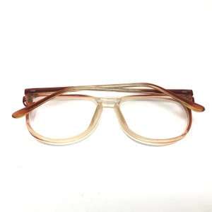 May include: Clear-framed eyeglasses with a gradient tint, transitioning from a light peach to clear. The temples are a darker brown with a thin, metallic accent. The glasses are set against a white background.