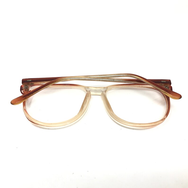 May include: Clear-framed eyeglasses with a gradient tint, transitioning from a light peach to clear. The temples are a darker brown with a thin, metallic accent. The glasses are set against a white background.