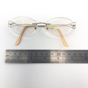 May include: Rimless eyeglasses with oval lenses and a silver-tone metal frame. The temples are a light peach color. A ruler is placed below the glasses, measuring approximately 6 inches in length.