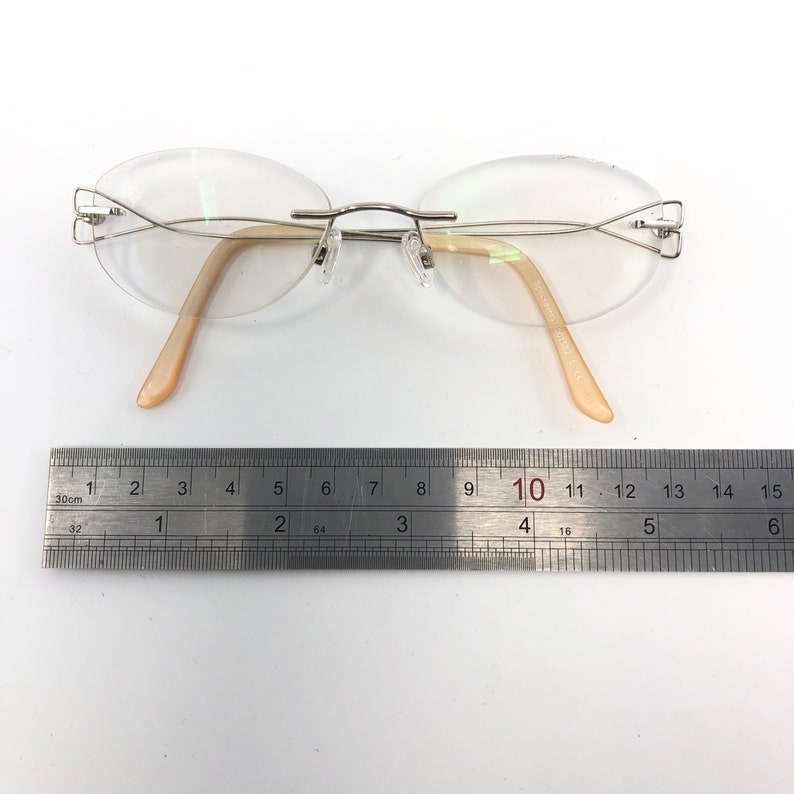 May include: Rimless eyeglasses with oval lenses and a silver-tone metal frame. The temples are a light peach color. A ruler is placed below the glasses, measuring approximately 6 inches in length.