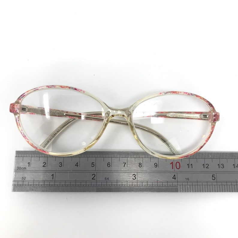 May include: Clear eyeglasses with oval lenses and a floral pattern on the frames. The frames are a light, translucent color with silver-toned arms. A ruler is placed below the glasses, showing measurements in inches.