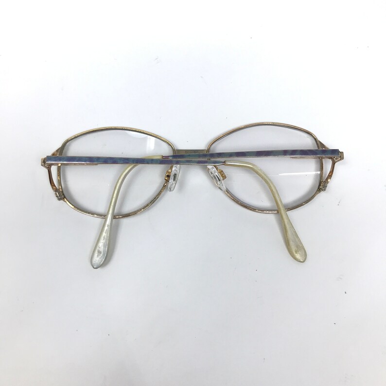 Op de afbeelding: Een bril met een goudkleurig metalen montuur en heldere glazen. De bril heeft een rechthoekige vorm met afgeronde randen. De veren zijn licht goudkleurig en de neuspads zijn transparant.