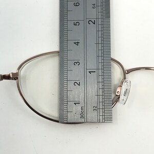 May include: Close-up of eyeglasses with a rose gold frame. A ruler is placed over the lens, measuring approximately 1 inch. The glasses have clear lenses and nose pads. The background is white.