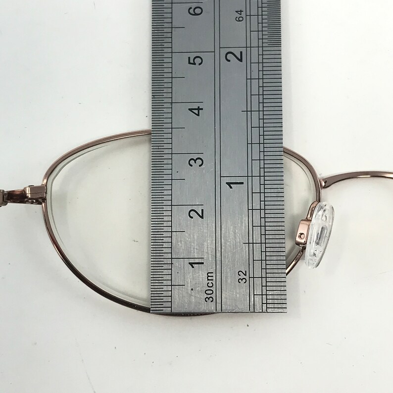 May include: Close-up of eyeglasses with a rose gold frame. A ruler is placed over the lens, measuring approximately 1 inch. The glasses have clear lenses and nose pads. The background is white.