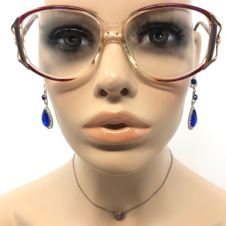K&ouml;nnte beinhalten: Brille mit transparentem Rahmen und burgunderroten und goldenen Akzenten am oberen Rahmen. Die Brille wird mit baumelnden Ohrringen mit blauen tropfenf&ouml;rmigen Edelsteinen und einer silbernen Halskette mit Herzanh&auml;nger kombiniert. Die Brille wird auf einer Schaufensterpuppe pr&auml;sentiert.