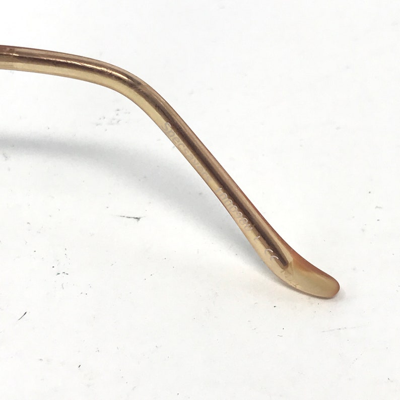 May include: Close-up of a gold-colored eyeglass arm. The arm curves gently and has the text "Specs" and "1000388 L" printed on it. The arm is smooth and has a slight sheen.