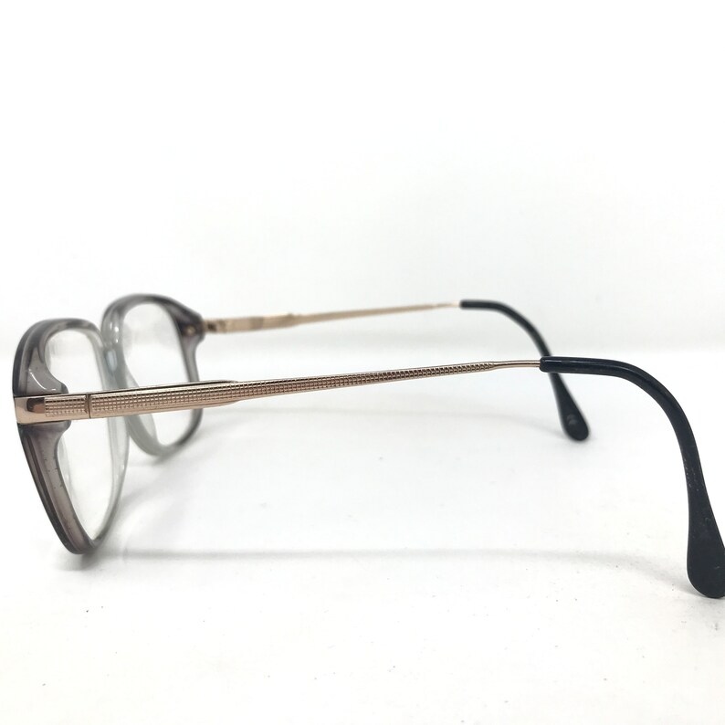 May include: Eyeglasses with a grey frame and clear lenses. The temples are gold-coloured with a textured design, and the earpieces are black. The glasses are set against a white background.