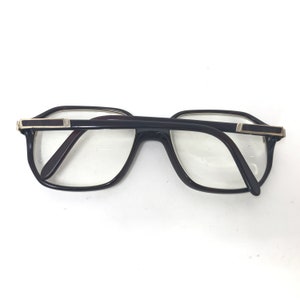 May include: Black-framed spectacles with a rectangular shape and clear lenses. The glasses have a dark brown interior and silver-coloured accents on the temples. The frames are thick and the lenses are large.