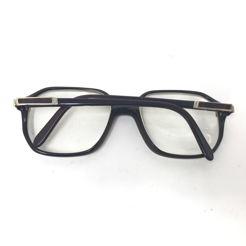 May include: Black-framed spectacles with a rectangular shape and clear lenses. The glasses have a dark brown interior and silver-coloured accents on the temples. The frames are thick and the lenses are large.