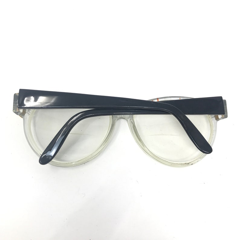 May include: Clear plastic framed glasses with black plastic arms. The glasses have a rounded shape.