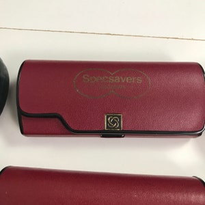 May include: A burgundy eyeglasses case with the Specsavers logo in gold. The case has a black trim and a gold clasp with the Specsavers logo. The case is rectangular and appears to be made of leather.