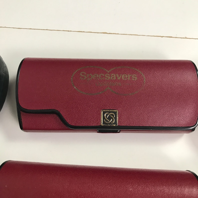 May include: A burgundy eyeglasses case with the Specsavers logo in gold. The case has a black trim and a gold clasp with the Specsavers logo. The case is rectangular and appears to be made of leather.