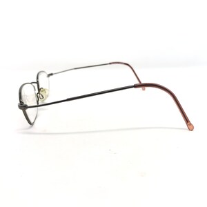 Puede incluir: Un par de gafas con montura fina y oscura y lentes ovaladas. Las patillas son de color marr&oacute;n rojizo, curv&aacute;ndose suavemente para adaptarse detr&aacute;s de las orejas. Las gafas est&aacute;n sobre un fondo blanco liso.