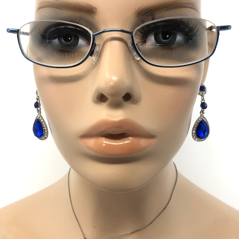 May include: A mannequin head wearing blue-rimmed eyeglasses and dangling earrings. The earrings feature large, teardrop-shaped blue gems surrounded by small, clear crystals. A silver necklace is also visible.