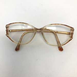 May include: Clear eyeglasses with a light brown frame and gold-colored arms. The lenses are oval-shaped, and the frame has a decorative pattern on the edges. The glasses are set against a white background.