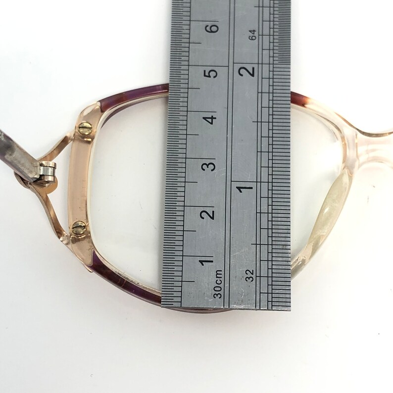 K&ouml;nnte beinhalten: Vintage Brille mit transparentem Rahmen und lila Akzenten. Ein Metalllineal wird neben die Brille gelegt und zeigt ein Ma&szlig; von etwa 3,8 cm. Die Brille hat eine rechteckige Form und ein goldfarbenes Metallscharnier.