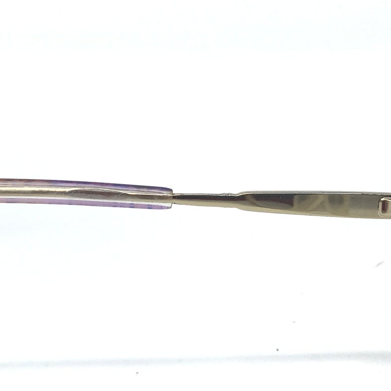 May include: Close-up of eyeglasses with a silver metal frame and purple temples. The glasses have a sleek, modern design, with a focus on the details of the frame and the temples. The image is set against a white background.