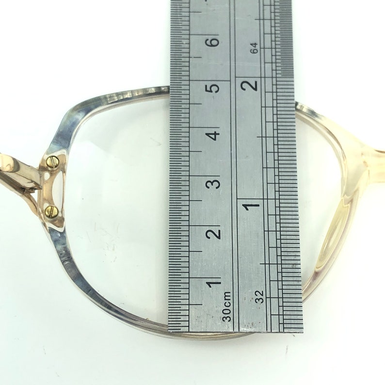 May include: Close-up of a pair of eyeglasses with a ruler measuring the frame. The glasses have a silver and gold frame with clear lenses. The ruler shows a measurement of approximately 2 inches.