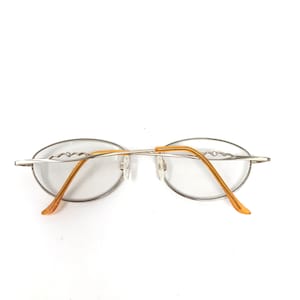 Puede incluir: Gafas de forma ovalada con montura plateada y varillas doradas. Las gafas tienen un dise&ntilde;o minimalista, con las lentes sujetas por una fina montura met&aacute;lica. Las varillas se curvan suavemente y terminan en color marr&oacute;n claro.