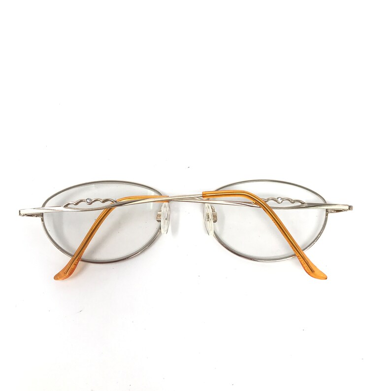 Puede incluir: Gafas de forma ovalada con montura plateada y varillas doradas. Las gafas tienen un dise&ntilde;o minimalista, con las lentes sujetas por una fina montura met&aacute;lica. Las varillas se curvan suavemente y terminan en color marr&oacute;n claro.