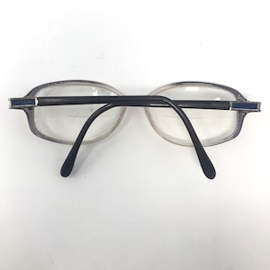 May include: A pair of eyeglasses with oval-shaped lenses and a black bridge and arms. The frames are a light purple color with a silver and blue accent on the temples. The lenses are clear.