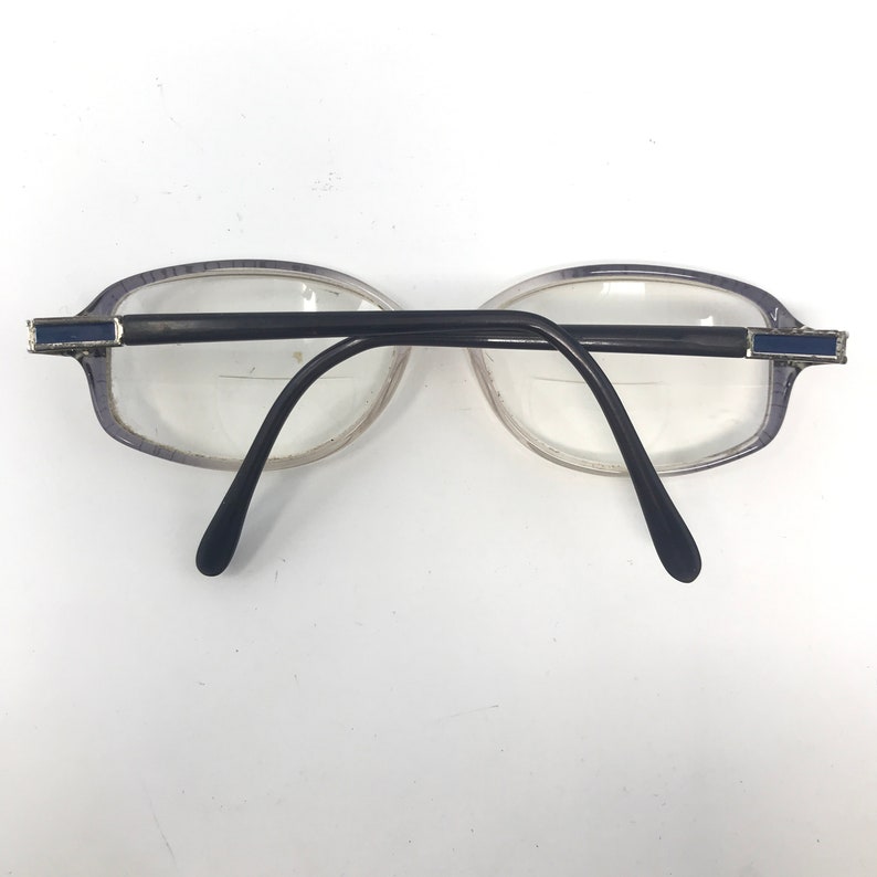 May include: A pair of eyeglasses with oval-shaped lenses and a black bridge and arms. The frames are a light purple color with a silver and blue accent on the temples. The lenses are clear.