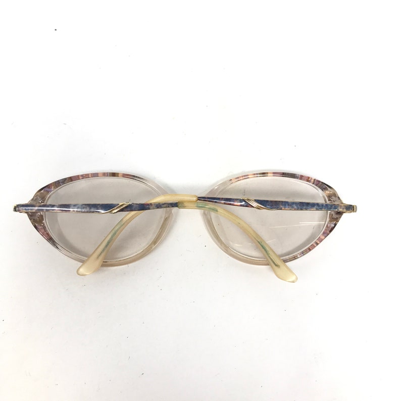May include: Vintage eyeglasses with oval lenses. The frames feature a marbled pattern in shades of pink, purple, and beige. The temples are a light cream color, and the bridge is a pale yellow. The metal accents are silver with gold detailing.