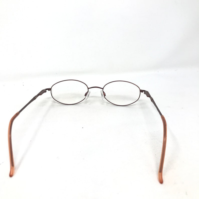 May include: Oval-shaped eyeglasses with a dark brown metal frame and clear lenses. The temples are a lighter brown color. The glasses are set against a white background.