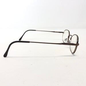 Puede incluir: Gafas de montura redonda con montura de metal color bronce y patillas negras. Las lentes son transparentes, sobre un fondo blanco. El dise&ntilde;o es sencillo y cl&aacute;sico.