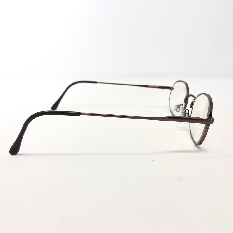 Puede incluir: Gafas de montura redonda con montura de metal color bronce y patillas negras. Las lentes son transparentes, sobre un fondo blanco. El dise&ntilde;o es sencillo y cl&aacute;sico.
