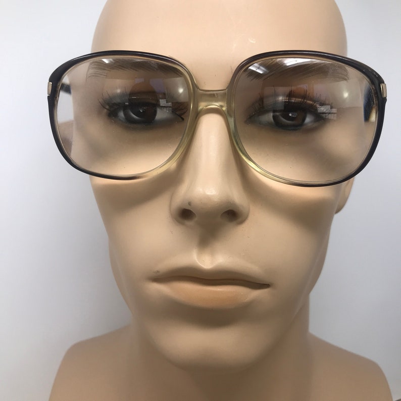 May include: Square-framed eyeglasses with a brown and clear plastic frame. The lenses are clear, and the glasses are displayed on a mannequin head. The glasses have a retro, vintage style.