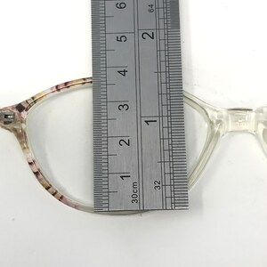 May include: Clear eyeglasses with a floral pattern on the frame are measured with a ruler. The ruler shows the glasses are approximately 1.5 inches wide. The glasses have a clear bridge and arms.