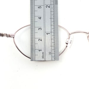 Puede incluir: Primer plano de unas gafas con montura plateada. Una regla se coloca sobre la lente, midiendo aproximadamente 2,5 cm. Las gafas tienen una forma ovalada cl&aacute;sica y lentes transparentes. El fondo es blanco.