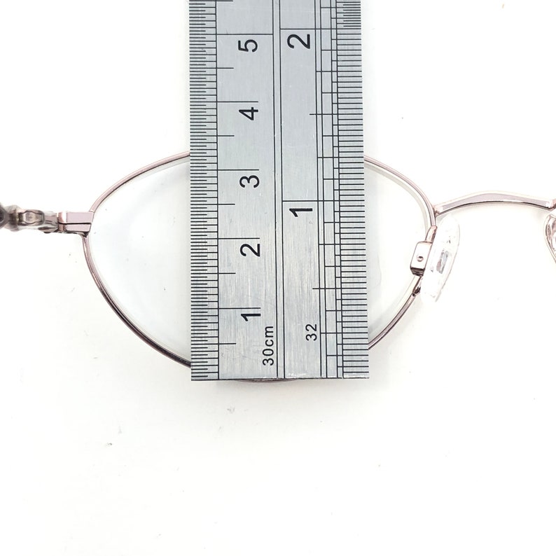 Puede incluir: Primer plano de unas gafas con montura plateada. Una regla se coloca sobre la lente, midiendo aproximadamente 2,5 cm. Las gafas tienen una forma ovalada cl&aacute;sica y lentes transparentes. El fondo es blanco.