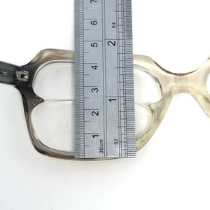 Op de afbeelding: Een bril met een transparant montuur en een grijze gradi&euml;nt op het bovenste montuur wordt gemeten met een liniaal. De liniaal toont dat de bril ongeveer 7,6 cm breed is. De bril heeft een unieke, hoekige vorm.