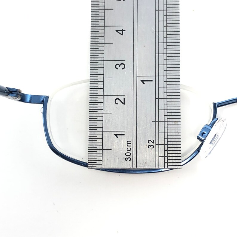 May include: Close-up of blue rectangular eyeglasses with a metal ruler placed over the lens. The ruler shows measurements in inches and centimeters. The glasses have a dark blue frame and clear lenses.