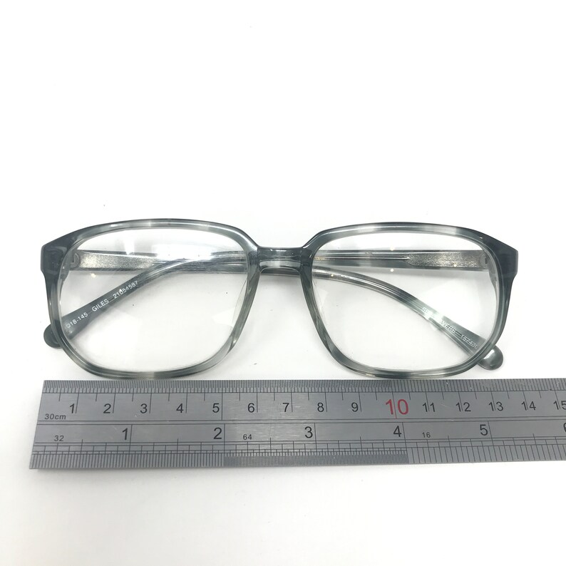 Puede incluir: Gafas grises de montura cuadrada con lentes transparentes. Las monturas tienen un patr&oacute;n veteado y un puente plateado. Una regla se coloca debajo, mostrando medidas en cent&iacute;metros. El texto "018-145 GILES 21854587" est&aacute; impreso en la montura.
