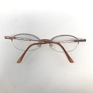 May include: Rimless eyeglasses with oval lenses and a brown metal frame. The temples and bridge are a matching brown color. The top of the frame has a decorative wavy design.