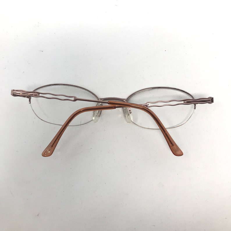 May include: Rimless eyeglasses with oval lenses and a brown metal frame. The temples and bridge are a matching brown color. The top of the frame has a decorative wavy design.