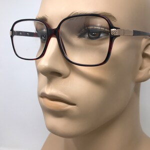 May include: Square-framed eyeglasses with a tortoiseshell pattern on the rims and temples. The glasses have a gold-colored accent on the temples and clear lenses. The frames are set against a neutral background.