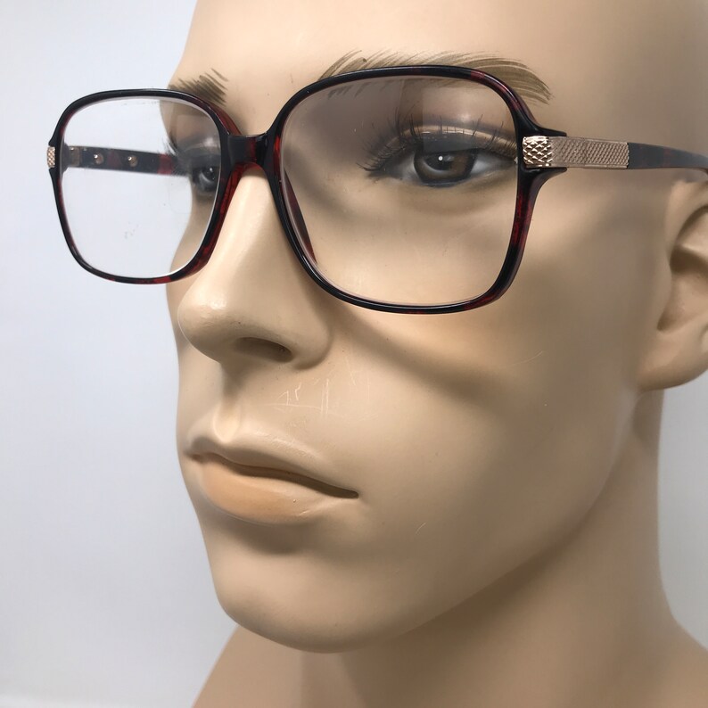 May include: Square-framed eyeglasses with a tortoiseshell pattern on the rims and temples. The glasses have a gold-colored accent on the temples and clear lenses. The frames are set against a neutral background.