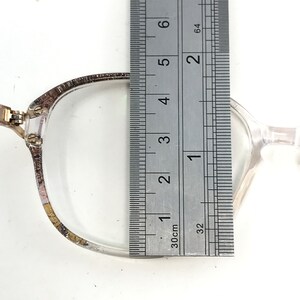 May include: Clear eyeglasses with a silver frame are measured with a ruler. The ruler shows the glasses are approximately 2 inches wide. The glasses are on a white surface.