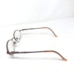 May include: Brown oval-shaped eyeglasses with clear lenses. The frames are a dark brown color, with a thin, delicate design. The temples have a lighter brown color at the ends. The glasses are set against a white background.