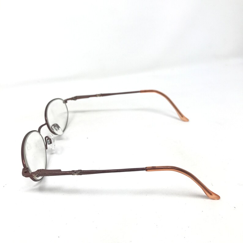 May include: Brown oval-shaped eyeglasses with clear lenses. The frames are a dark brown color, with a thin, delicate design. The temples have a lighter brown color at the ends. The glasses are set against a white background.
