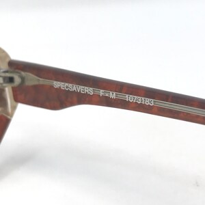 May include: Close-up of a brown and tan speckled eyeglass frame arm. The text "SPECSAVERS F-M 1073183" is printed on the arm in a silver color. The frame arm is made of a speckled brown material.
